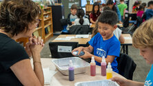 boy smiling as he uses spray bottle filled with color liquid as teacher looks on